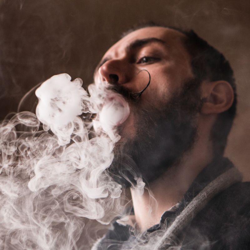 Young Man with Beard and Mustages Vaping an Electronic Cigarette. Vaper Hipster Smoke Vaporizer and Exhals Smoke Rings.